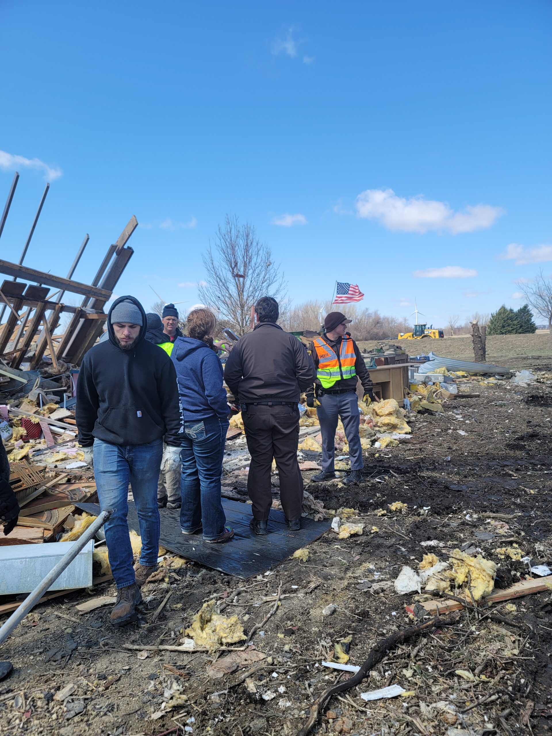 Disaster scene with people walking among strewn insulation, pipes and wall sections, US flag in the background