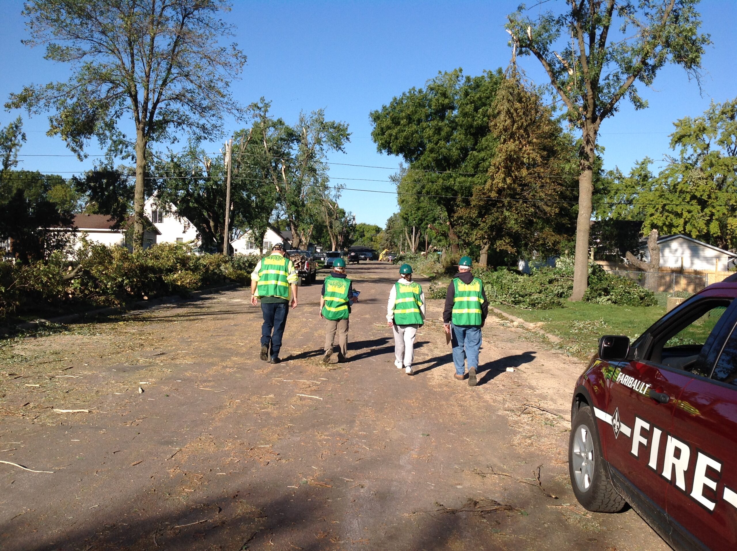 Four people in safety vests walking away from a Fire vehicle down a road littered with fallen trees.